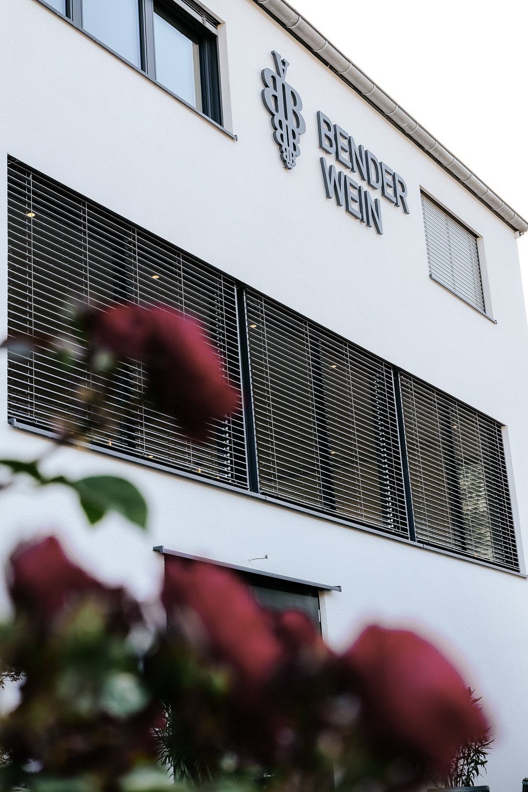Ein hohes, weißes Gebäude mit einem Schild an der Fassade, im Vordergrund von rosa Blumen umgeben.