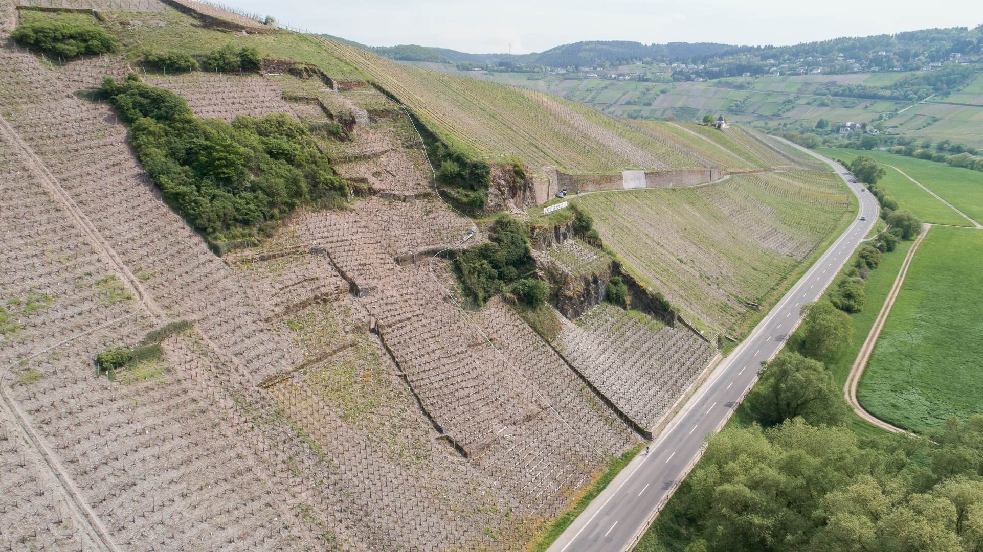 Luftaufnahme einer hügeligen Landschaft mit terrassierten Flächen und Vegetation, umgeben von sanften Hügeln.
